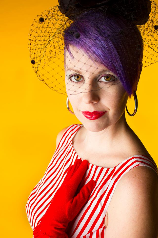 portrait of Roxanne Abell with a bright mustard yellow background cropped at the breasts. Her hair is short purple and purple under a black 1950s style hat she is wearing with the netting covering her hazel eyes. Large silver hoop earrings dangle from her mostly hidden ears. Her lips are red like the glove on her left hand that is laying upon her chest where her heart would be. She is in a red and white vertical striped sleeveless boatneck dress. There are freckles on the peachy/tan skin of her left shoulder which is exposed to the camera. She is looking directly at the camera with an engaging, and content expression. Her closed red lipped mouth turned slightly up in a smile. 